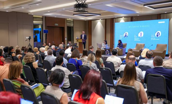 The image shows a conference.A speaker stands at a podium addressing a seated audience, while three panelists are seated on a stage with a large screen behind them. The screen displays the event's title, "Welcoming Remarks," along with the names and photos of the speakers. The audience, composed of men and women, is seated in rows, attentively listening and taking notes on laptops. 