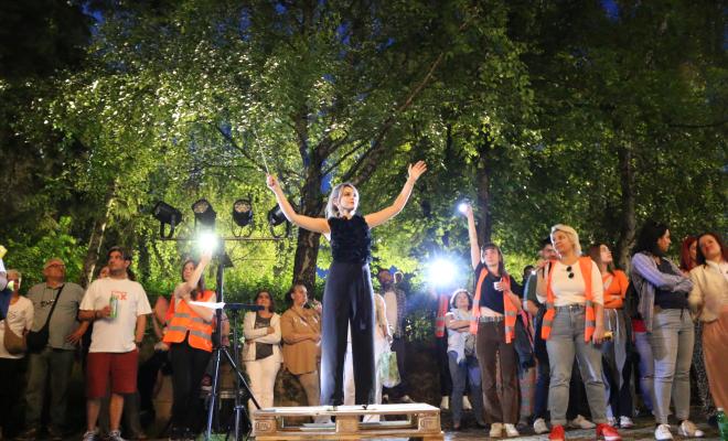 In the image, a performer stands on a wooden pallet, arms raised, engaging the audience at a festival in the Zelezara neighborhood. The event takes place outdoors, with people watching attentively.